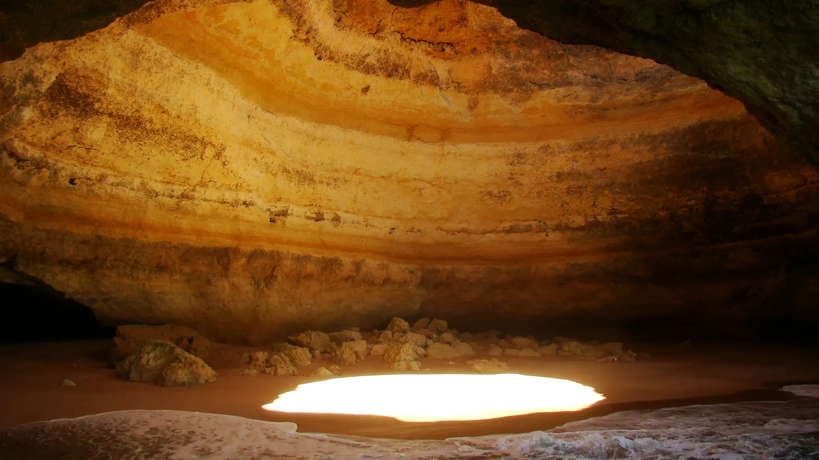 Das Innere der Benagil-Höhle: geschichteter ockerfarbener Fels und der Lichtfleck des Deckenlochs auf dem Sand